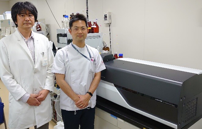 Photo: Associate Professor Takahiro Kato (left), and Assistant Professor Daiki Setoyama of Clinical Laboratories at Kyushu University Hospital, and the High-Performance LC-MS Used in the Research
