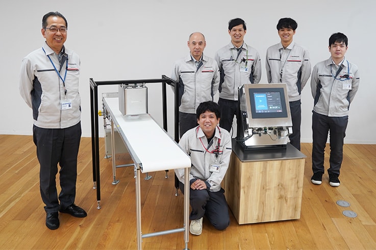 Front row center: Satoshi Kuno, who led the development. Back row, from left: Naoki Yoshioka, Atsufumi Ogishi, Soichiro Kitamura, Sogo Miura, Haruhiko Sakai. The equipment above the platform simulating a production line is the MMS series